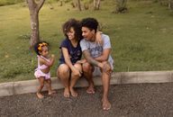 Cheerful African American mother and father with cute little daughter on kerb near lawn with verdant grass in daytime