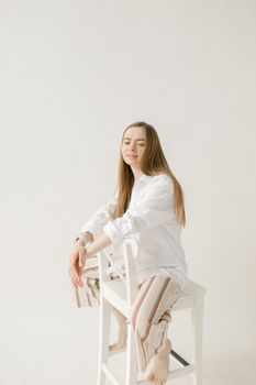 A young woman with long hair sits on a chair in a minimalist studio environment wearing a white shirt and striped pants.