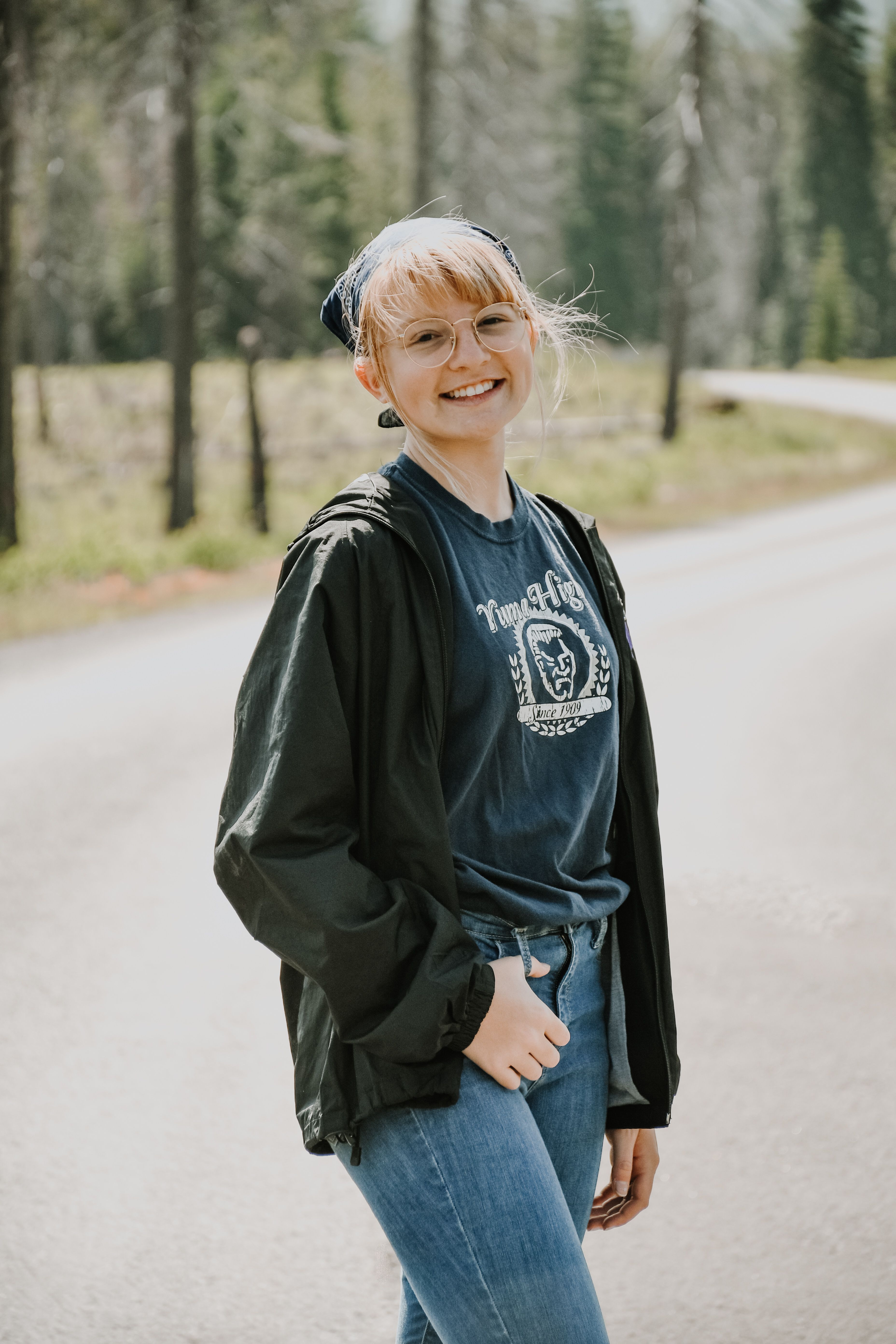 A young woman in casual outfit smiling warmly on a sunlit road surrounded by nature.