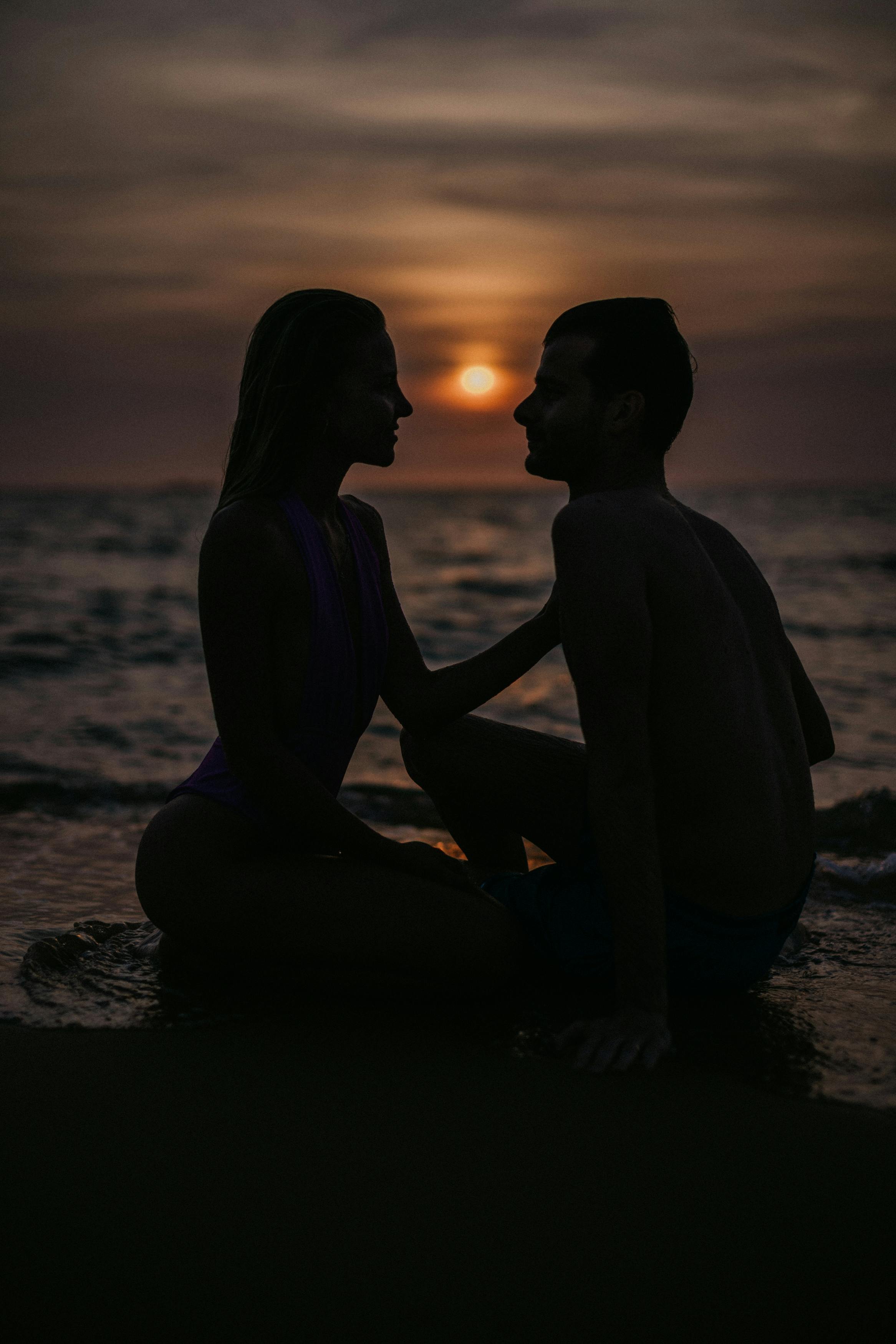 silhouette of a couple looking at each other while sitting on the beach during sunset