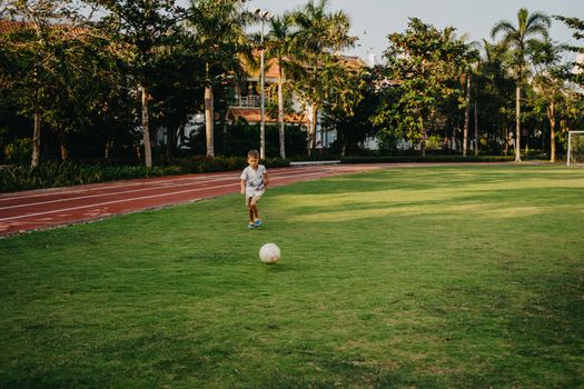A young boy plays soccer on a sunny field, surrounded by trees and a running track.