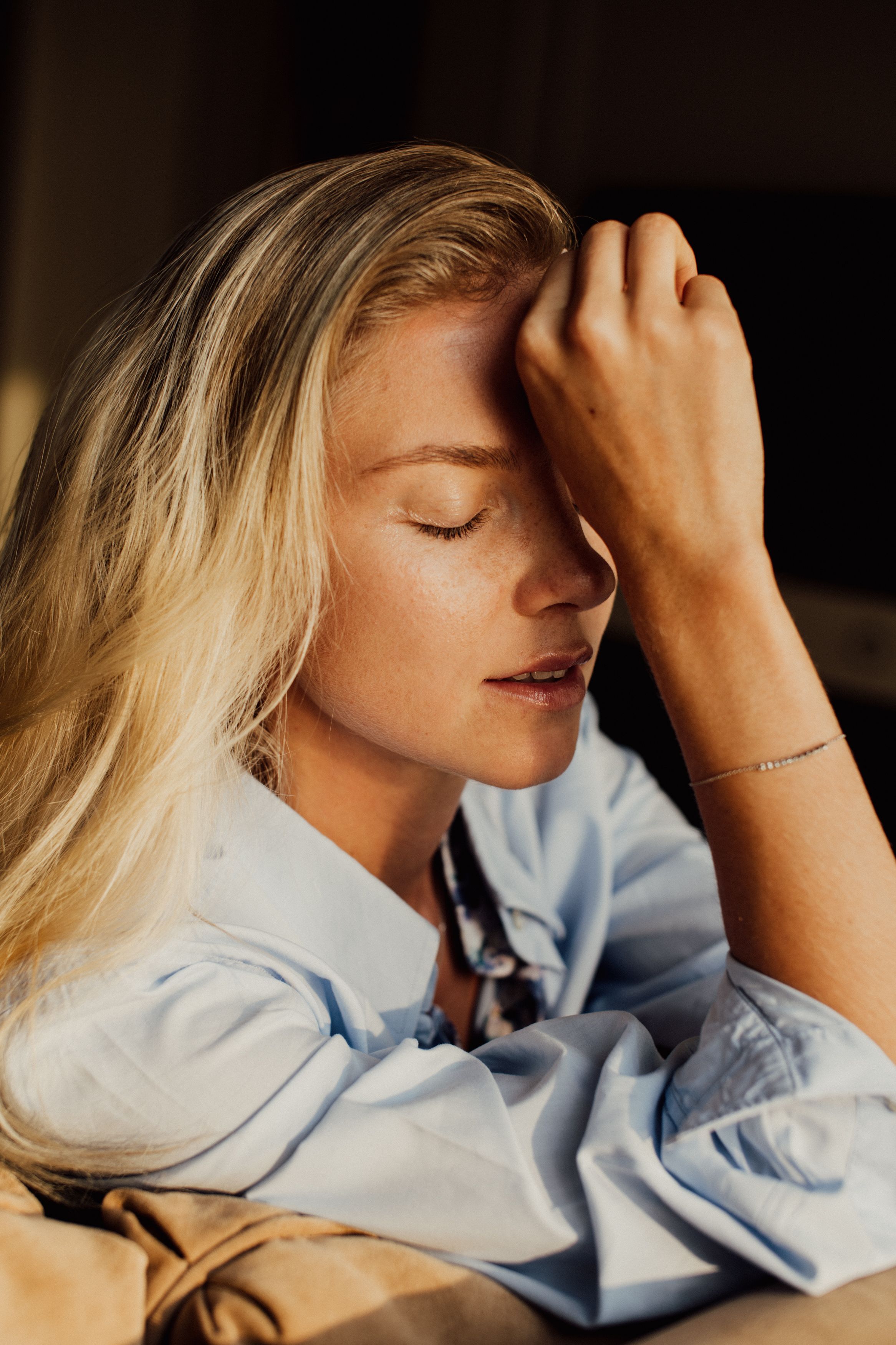 A serene close-up of a blonde woman with her hand on her head, bathed in gentle indoor lighting.