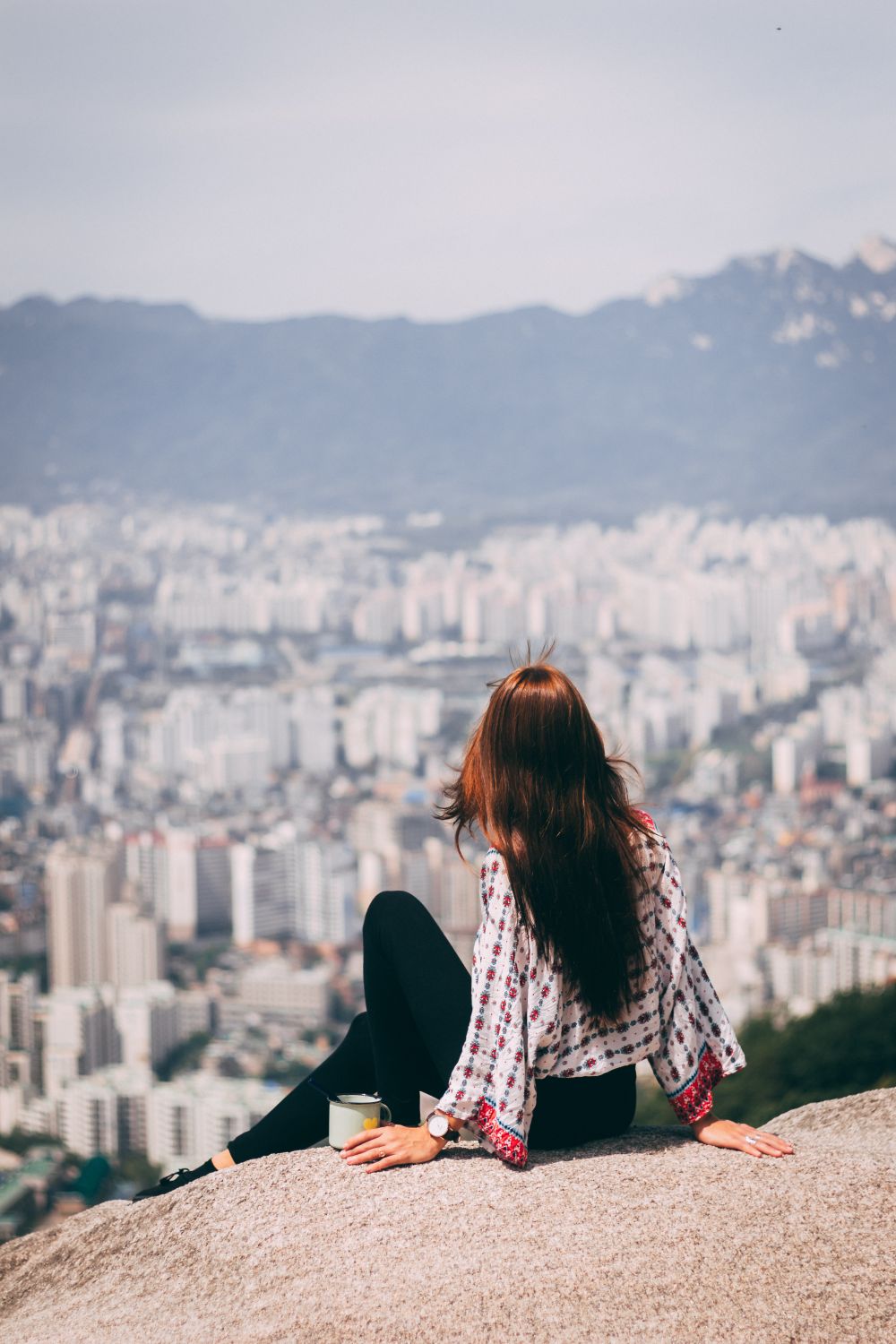 Woman sitting on rock overlooking Seoul urban landscape from a hillside viewpoint