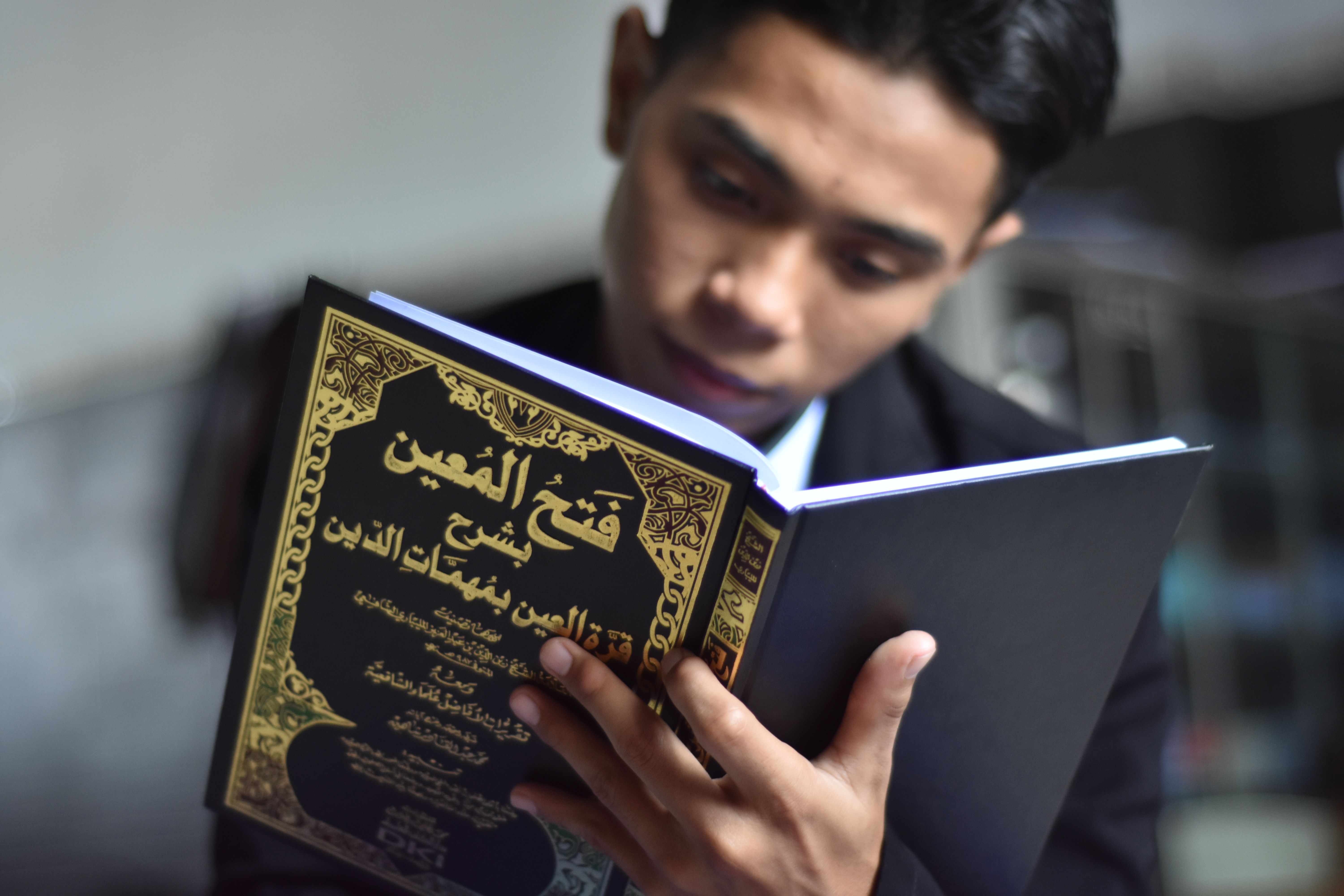 A young man intently reads an Islamic book indoors, showcasing cultural engagement.