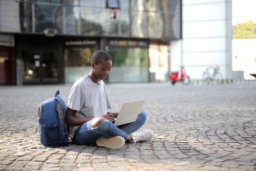 Free Teenager focuses on a laptop while sitting on city pavement, near a backpack and building. Stock Photo