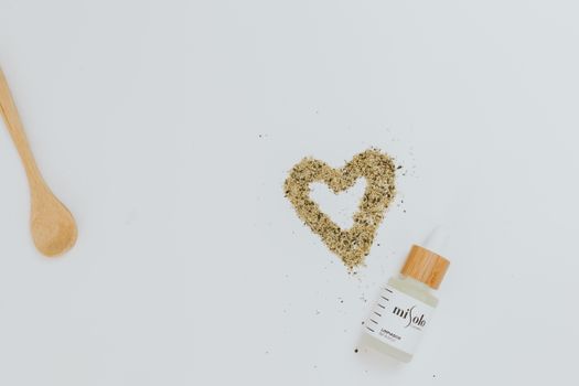 Flat lay of a cosmetic bottle next to a heart-shaped herbal design on a white background.