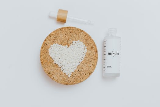 Serum bottle on white background with rice grains shaped as heart on cork board, emphasizing skincare and love.