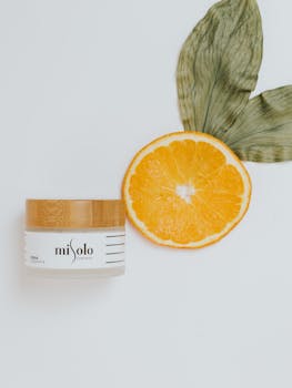 A minimalist display of a natural cosmetic jar with half an orange slice on a white background.