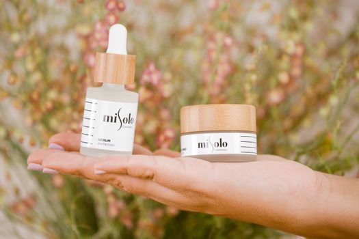 Close-up of hands holding natural cosmetic products outdoors with a floral background.