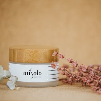 A beautifully styled cosmetic jar with pink flowers and a wooden lid on a beige background.