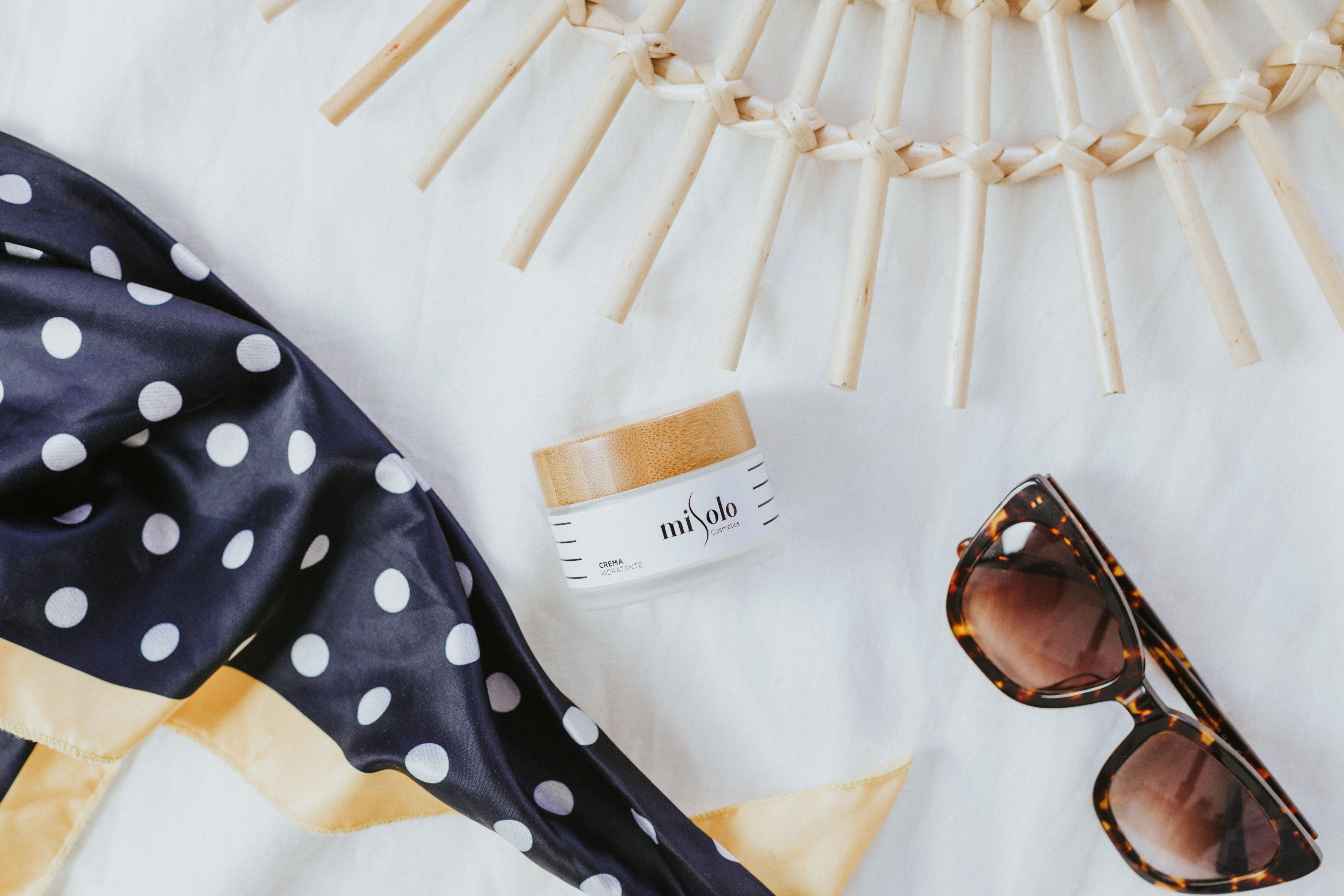 Chic flat lay featuring cosmetic cream, polka dot scarf, and sunglasses on a white surface.