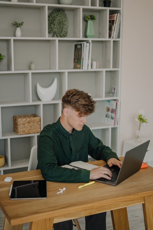 A Male Student in Green Long Sleeves Using a Laptop on a Wooden Desk