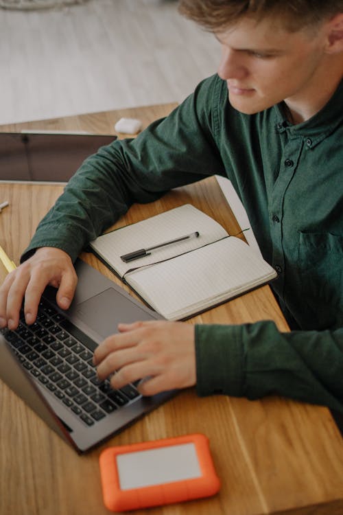 Free Close-Up Shot of a Male Student in Green Long Sleeves Using a Laptop on a Wooden Desk Stock Photo