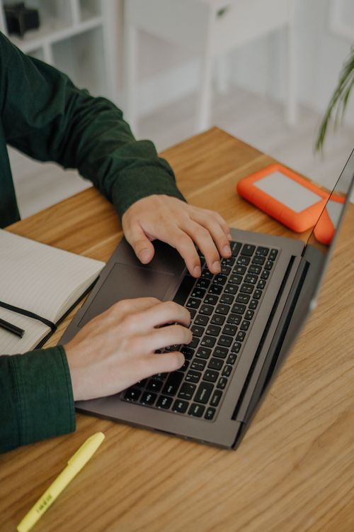 Free Hands typing on a laptop at a wooden desk with an external hard drive nearby. Stock Photo