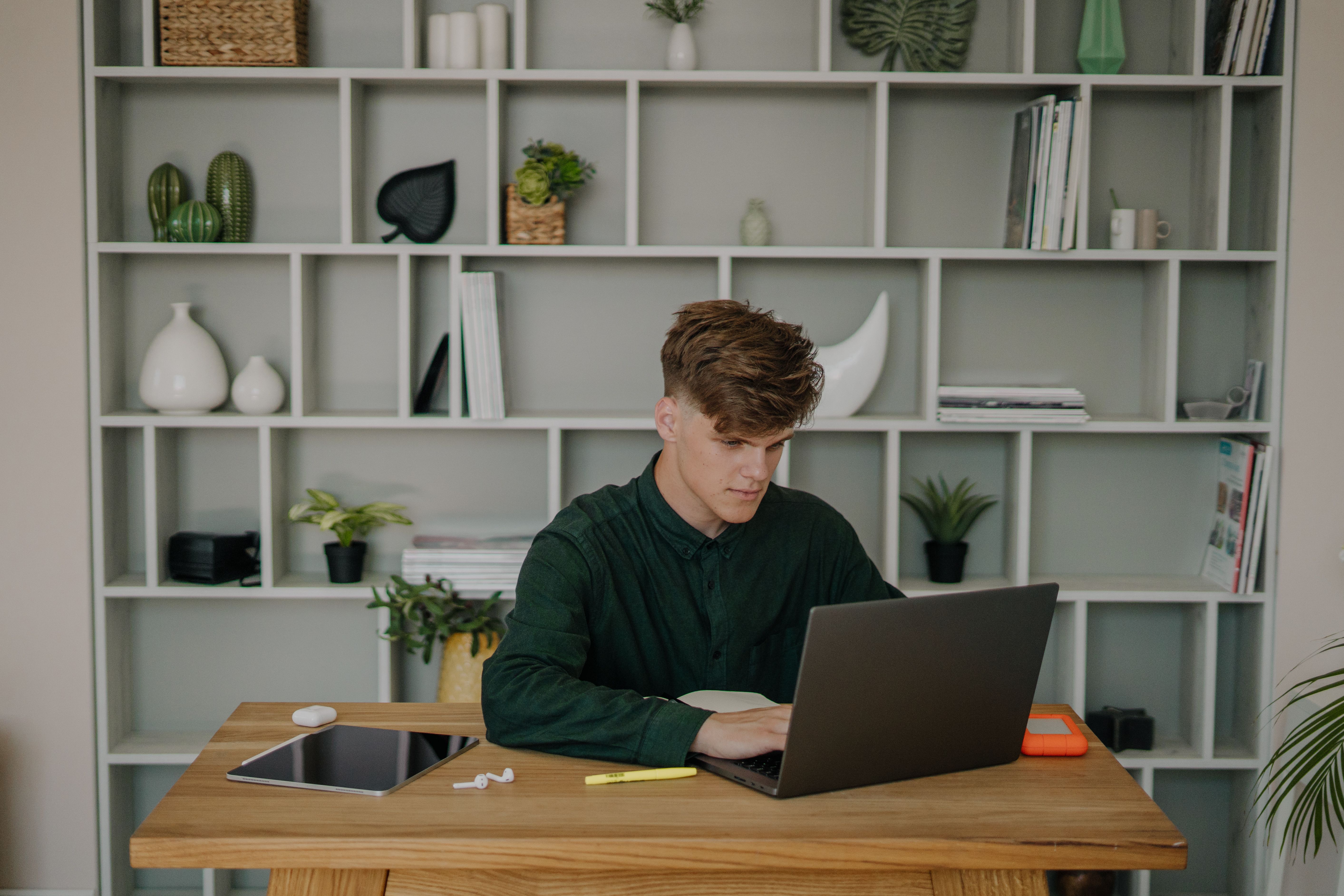 A man focused on using his laptop at a stylish home office desk with shelves.