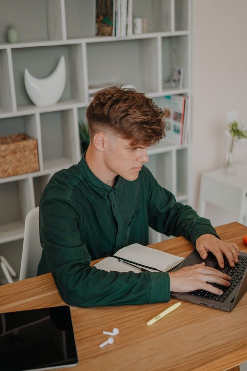Free A young man in a green shirt uses his laptop while studying at home in a modern setting. Stock Photo