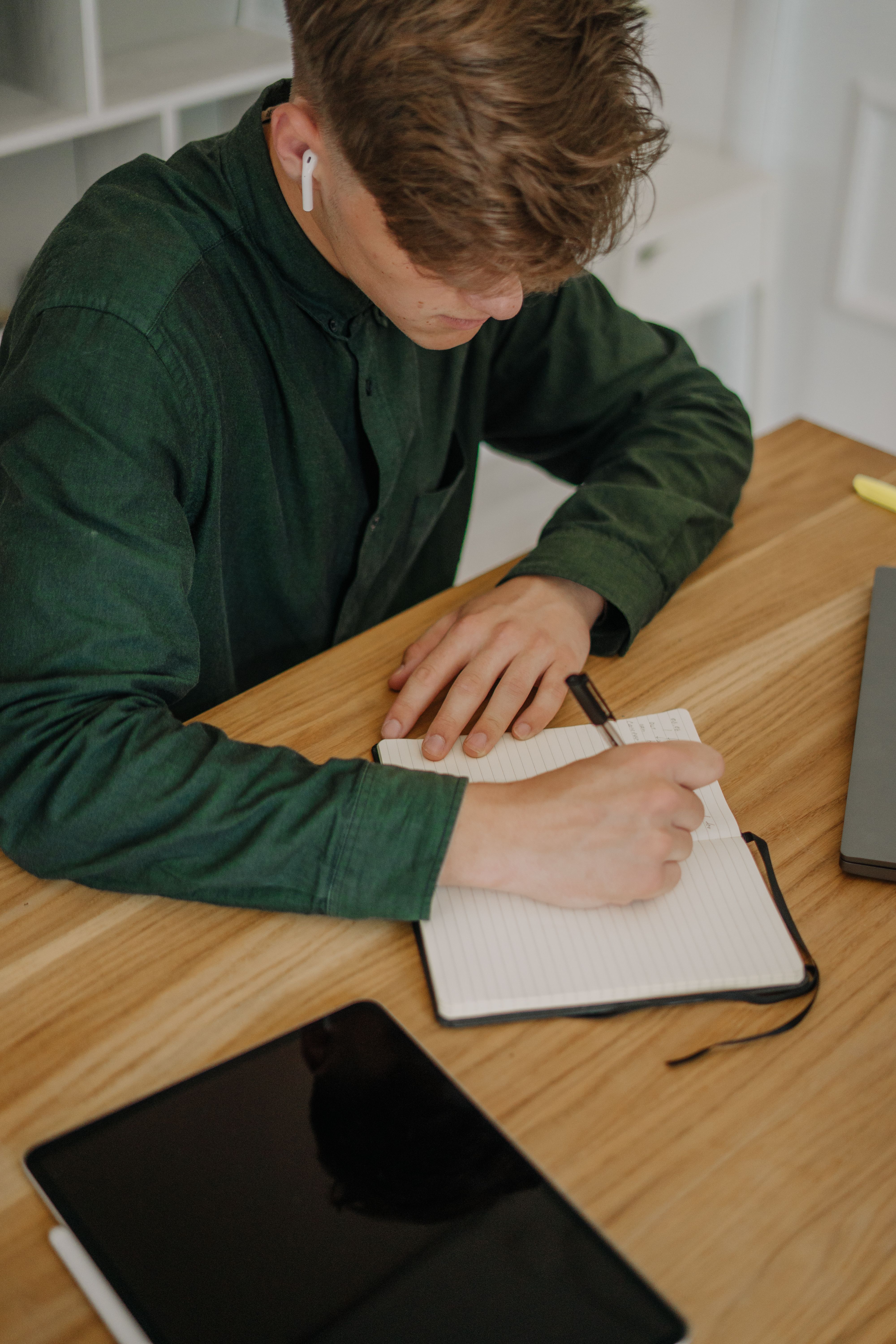 A young man in green shirt writes in a notebook while seated at a wooden desk, wearing earphones.