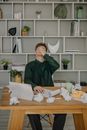 一名男子坐在办公桌前，被工作压得不堪重负，周围散落着皱巴巴的纸张和平板电脑