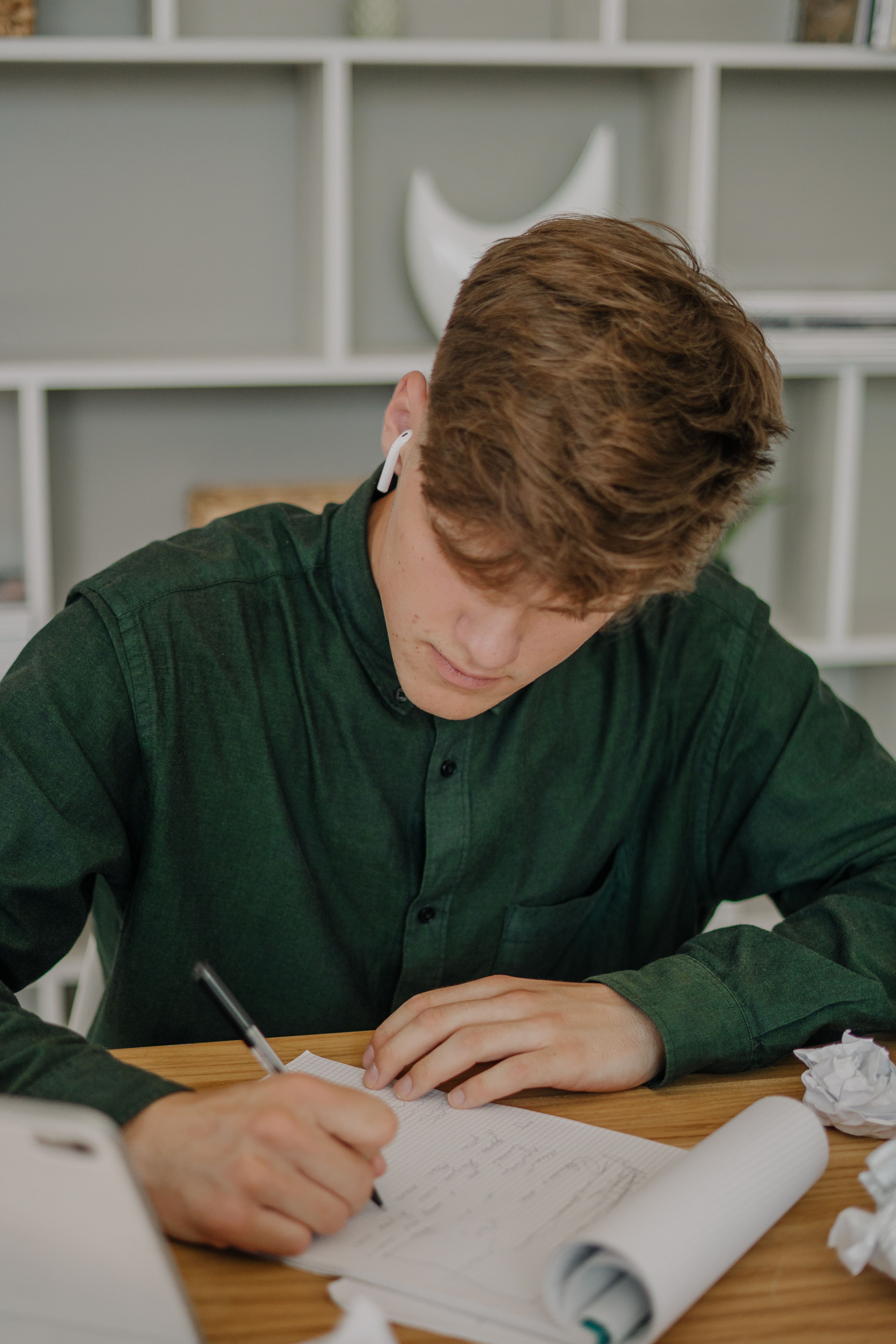 Young man in green shirt focused on taking notes at a wooden desk with earbuds in.