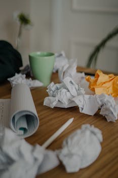 A cluttered office desk showcasing crumpled papers, a green coffee cup, and a pencil, evoking a creative yet chaotic workspace.