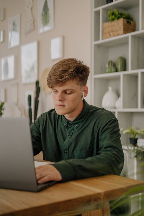 Close-Up Shot of A Male Student in Green Long Sleeves Using a Laptop on a Wooden Desk