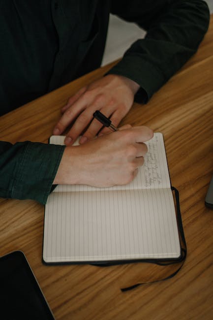 A person taking notes in a notebook with a pen on a wooden desk indoors.