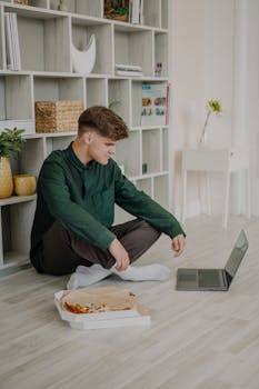 Young man sitting on the floor with laptop, eating pizza in a home office setting.