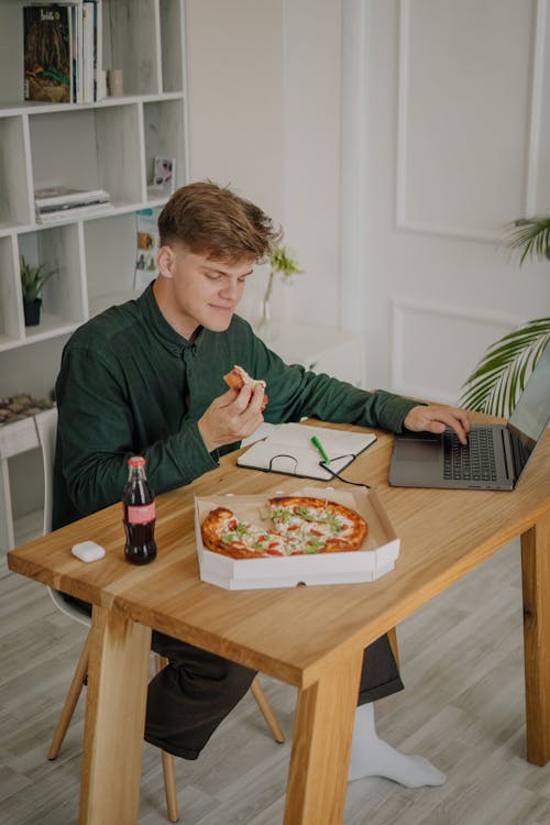 Young Man Eating Pizza and Working on His Laptop