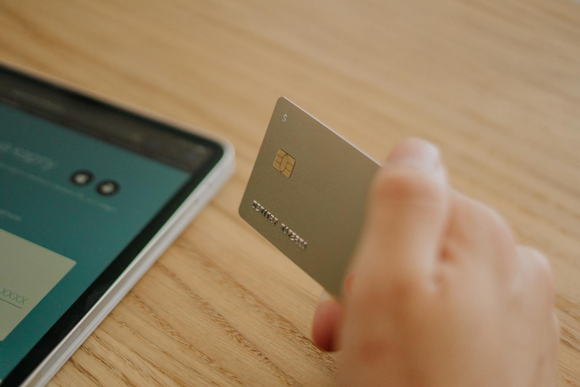 Close-up of a hand holding a credit card near a tablet, depicting online shopping or transaction.