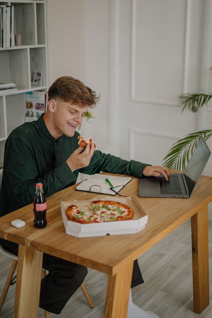 A man with green sleeves enjoying pizza and soda while working on a laptop at home.