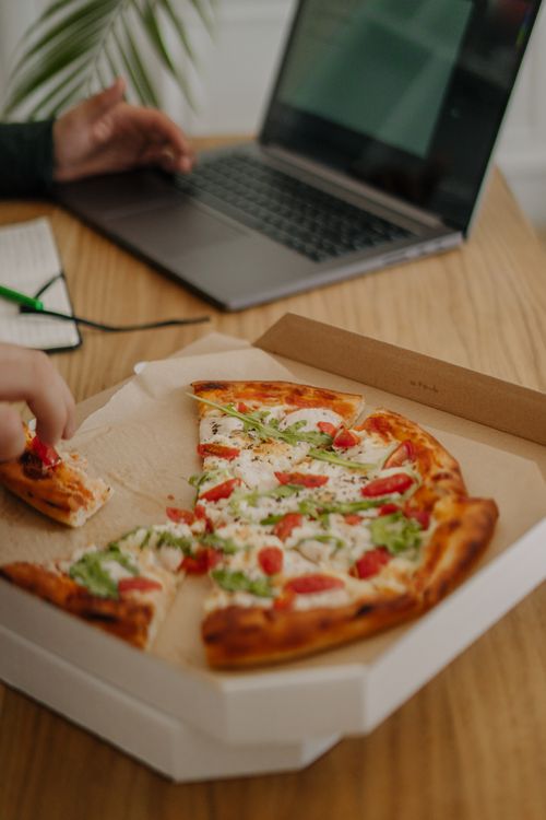 Free A person enjoys pizza while working on a laptop during lunchtime in an office setting. Stock Photo