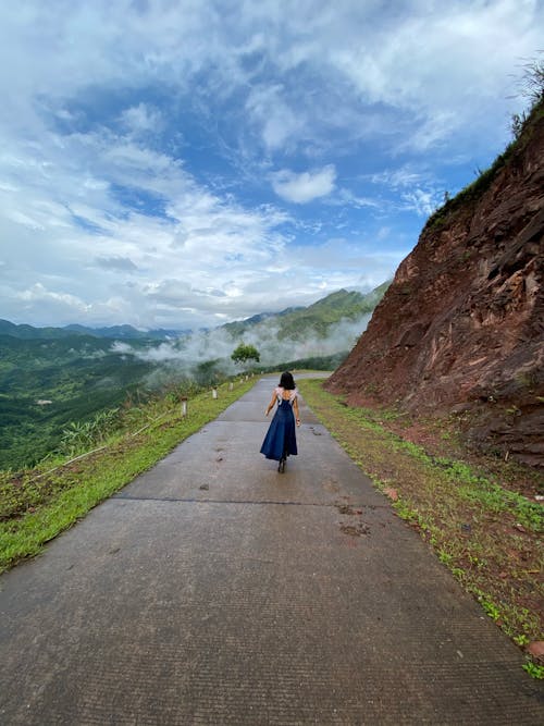 Back view of anonymous female in dress strolling on rural road along rocky cliff in highlands covered with fog against cloudy sky in nature
