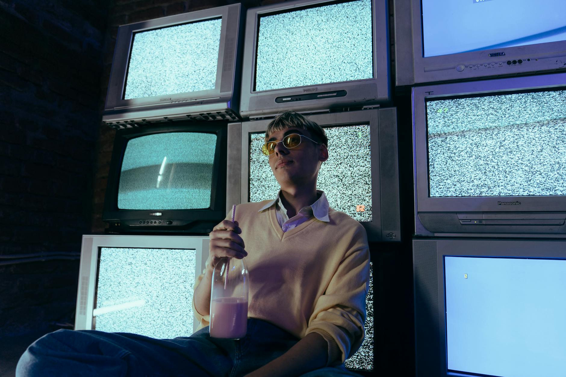 Teenage boy holding a milkshake surrounded by vintage CRT televisions showing static in a dimly lit room.