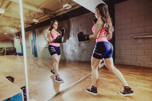 Back view concentrated sportswoman with perfect body wearing sportswear and boxing gloves punching air while looking in big wall mirror in modern gym