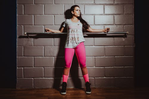 Full body confident fit sportswoman wearing pink leggings standing with arms outstretched near metal bar in gym and looking away