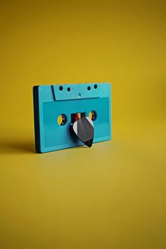 Vintage turquoise cassette tape holding a guitar pick on a vibrant yellow background.