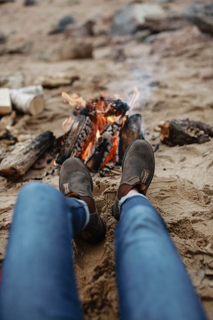 Close-up of legs in boots by a sandy beach campfire, cozy outdoor scene.