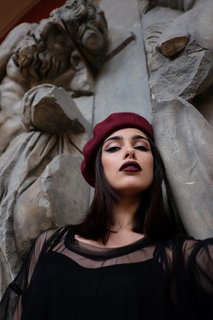 Low angle of young sensitive female in elegant wear and beret looking at camera near stone sculpture in museum