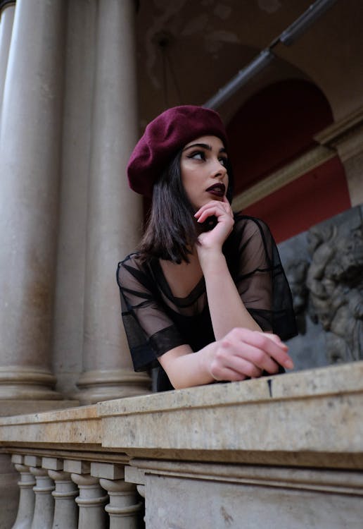Free Low angle of young contemplative elegant woman in beret touching chin while leaning with hand on masonry fence and looking away in old building Stock Photo