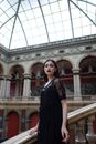 侧面图中，一位化着深色妆容、身着黑色服装的年轻黑发女子，站在古老的拱形建筑大厅里，注视着镜头