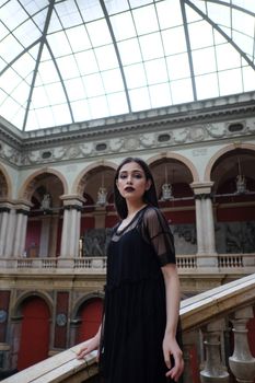 侧面图中，一位化着深色妆容、身着黑色服装的年轻黑发女子，站在古老的拱形建筑大厅里，注视着镜头