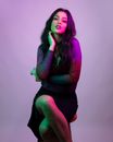 Modern female in trendy dress sitting on chair with legs crossed and hand on neck while looking at camera dreamily on purple background