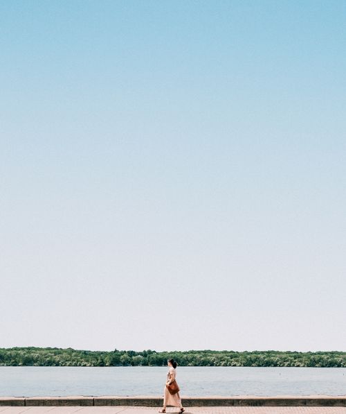Free A woman walks along a lakeside promenade on a clear, sunny day, with open skies and tranquil water. Stock Photo