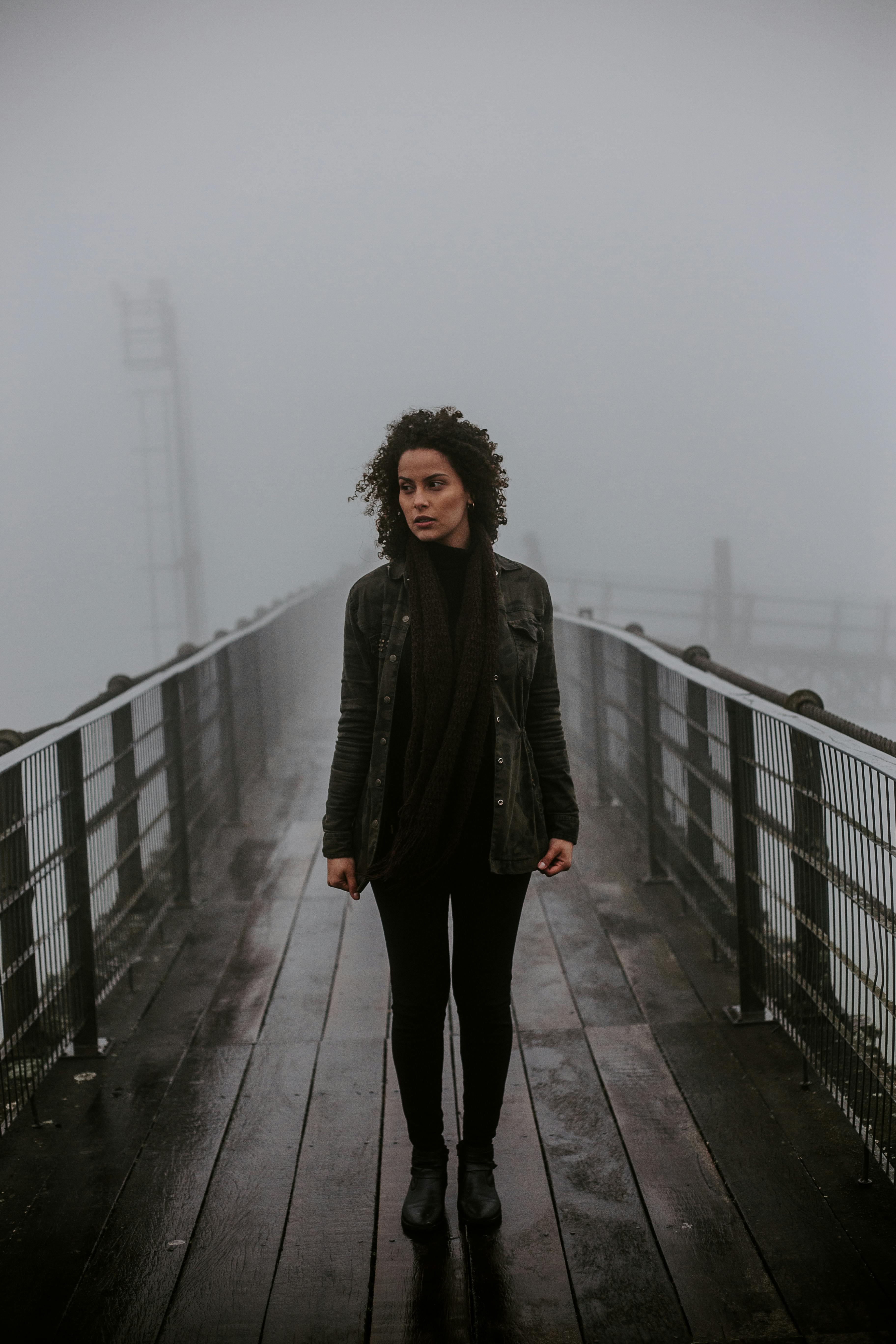 一位卷发女子穿着黑色服装，站在雾气缭绕的桥上，肖像喜怒无常