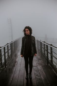一位卷发女子穿着黑色服装，站在雾气缭绕的桥上，肖像喜怒无常