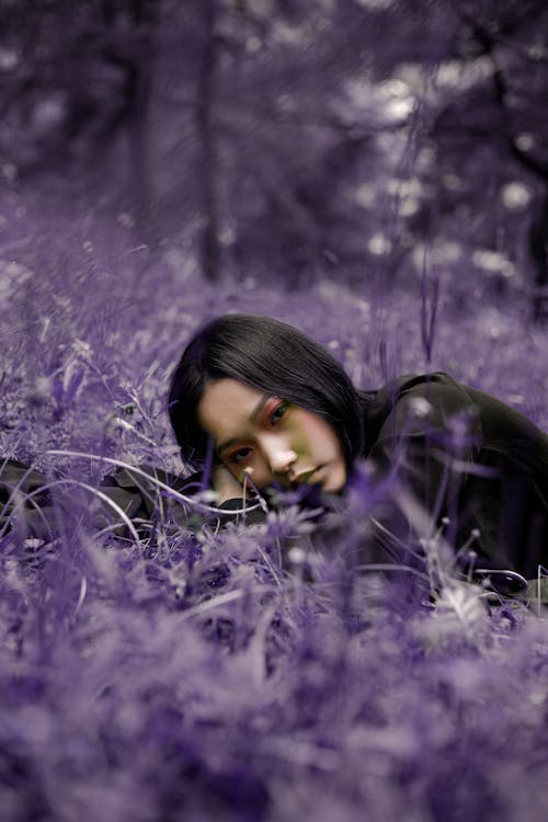 Young contemplative Asian female with makeup leaning with hand on blossoming violet flowers in daylight while looking at camera