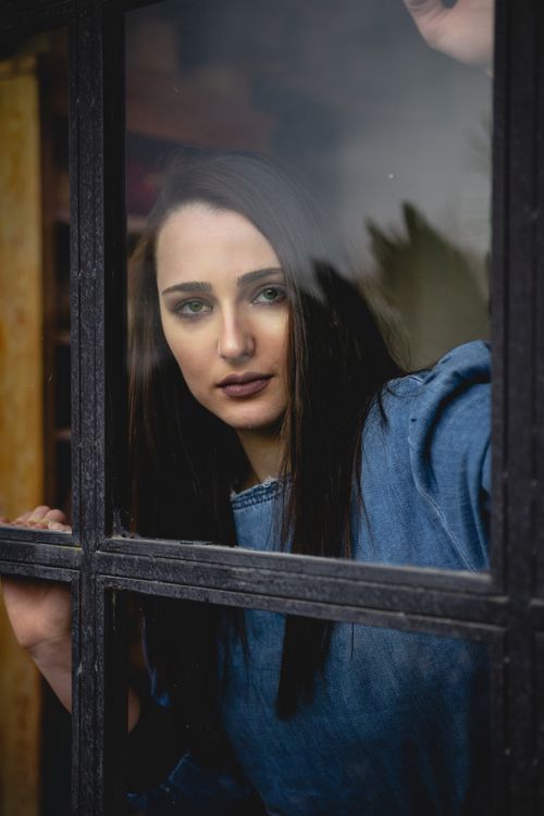 Free A thoughtful woman in a denim shirt looking through a window, capturing an introspective mood. Stock Photo
