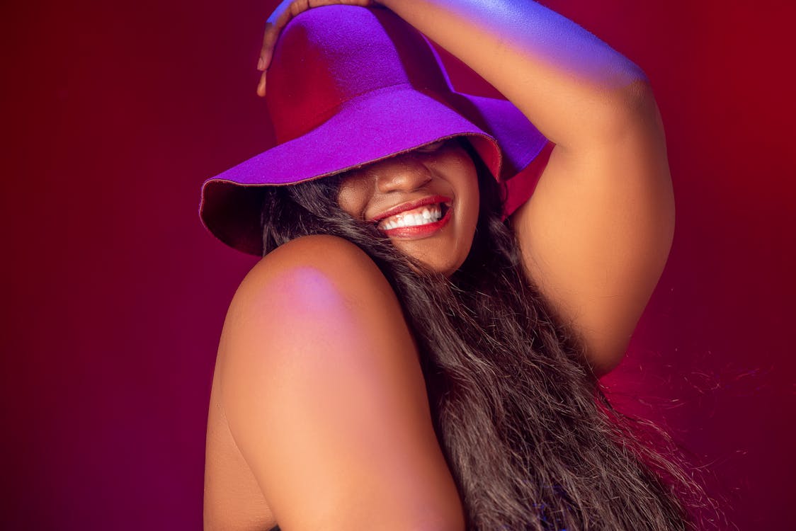 Free Side view of glad plus size African American female covering eyes with hat while holding hand on head and laughing Stock Photo