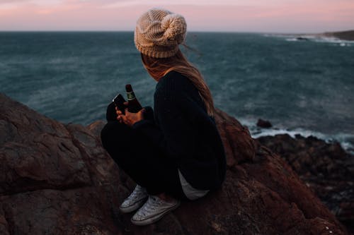 Anonymous young lady relaxing on rocky cliff and enjoying seascape under sundown sky