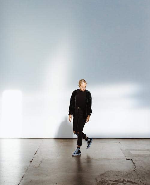 Full body of peaceful trendy young female millennial in stylish total black outfit and sneakers walking in spacious empty building against white wall